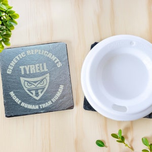 May include: Two square slate coasters with engraved text and a white coffee cup lid. One coaster reads "GENETIC REPLICANTS TYRELL MORE HUMAN THAN HUMAN" with an owl graphic. The other coaster is under the cup lid.