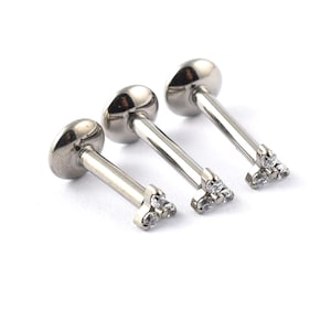 May include: Three silver lip piercing studs. Each stud has a flat, oval top and a straight post. The base of each post features three small, clear, sparkling stones. The studs are arranged diagonally against a white background.