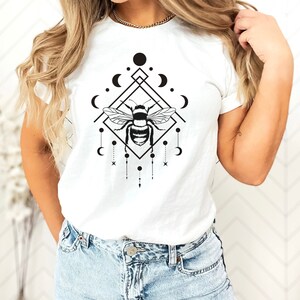 May include: White t-shirt featuring a black and white graphic design of a bee within a geometric shape, adorned with moons and stars.