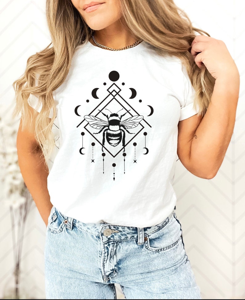 May include: White t-shirt featuring a black and white graphic design of a bee within a geometric shape, adorned with moons and stars.