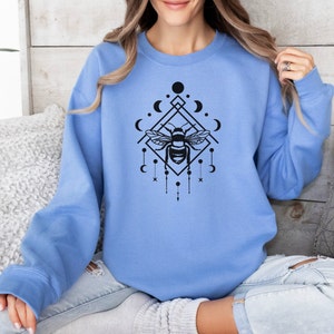 May include: A light blue sweatshirt featuring a black graphic of a bee within a geometric design incorporating moons and stars.