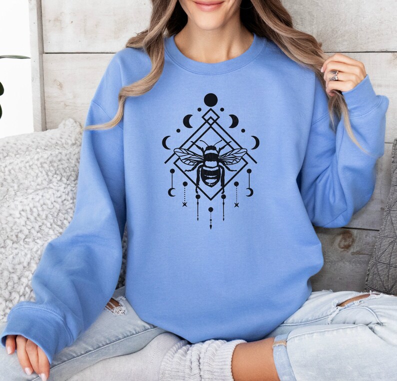 May include: A light blue sweatshirt featuring a black graphic of a bee within a geometric design incorporating moons and stars.