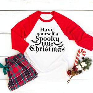 K&ouml;nnte beinhalten: Ein wei&szlig;es und rotes Baseball-T-Shirt mit dem Aufdruck "Have yourself a Spooky little Christmas" auf der Vorderseite. Das Shirt liegt auf einer wei&szlig;en Holzoberfl&auml;che mit einer roten und gr&uuml;nen karierten Schlafhose, einem Zuckerstangen und einem kleinen Weihnachtsbaum.