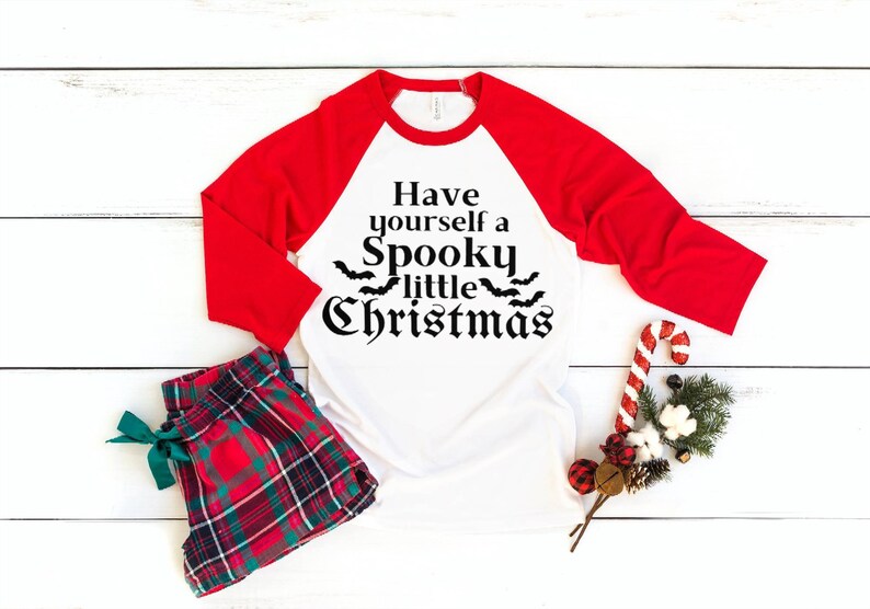 K&ouml;nnte beinhalten: Ein wei&szlig;es und rotes Baseball-T-Shirt mit dem Aufdruck "Have yourself a Spooky little Christmas" auf der Vorderseite. Das Shirt liegt auf einer wei&szlig;en Holzoberfl&auml;che mit einer roten und gr&uuml;nen karierten Schlafhose, einem Zuckerstangen und einem kleinen Weihnachtsbaum.