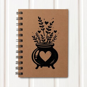 May include: A brown spiral bound notebook with a black illustration of a cauldron with a heart on the front. The cauldron is filled with black flowers and hearts.