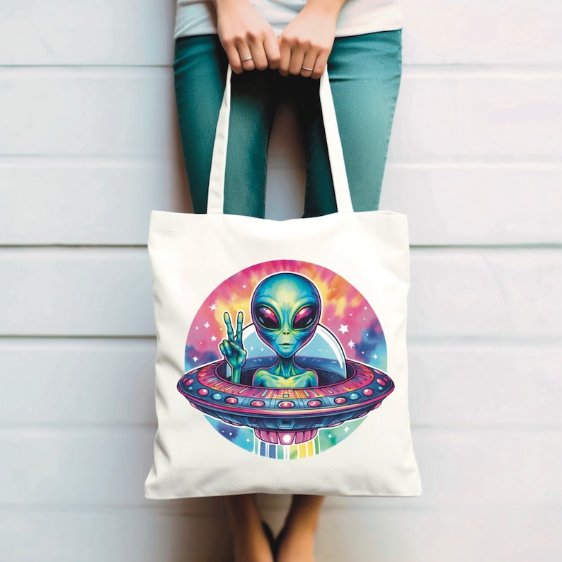 Puede incluir: Bolsa de tela blanca con un gr&aacute;fico colorido de un extraterrestre con piel verde y ojos grandes, haciendo un signo de paz, sentado en un platillo volante sobre un fondo de galaxia.