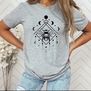 May include: A grey t-shirt featuring a black graphic of a bee within a geometric design incorporating moons and stars.