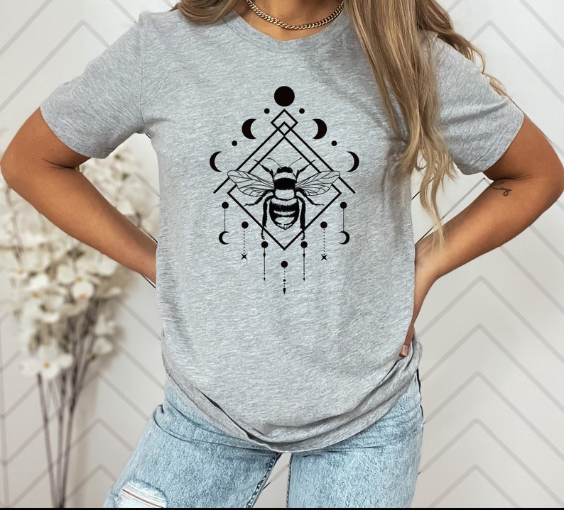May include: A grey t-shirt featuring a black graphic of a bee within a geometric design incorporating moons and stars.