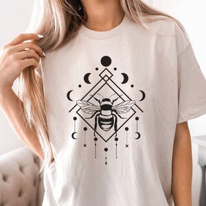 May include: A beige t-shirt featuring a black and white graphic design of a bee within a geometric shape, surrounded by moons and stars.