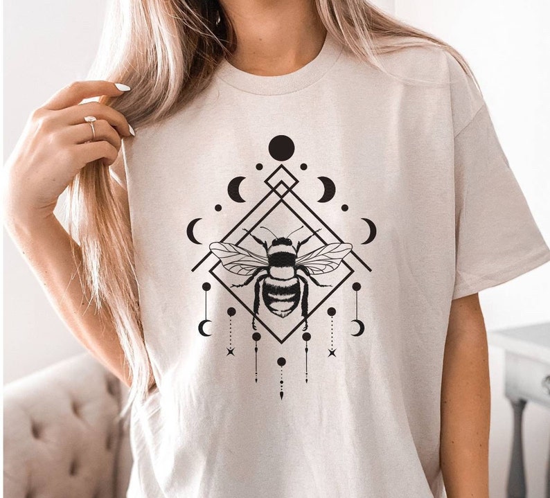 May include: A beige t-shirt featuring a black and white graphic design of a bee within a geometric shape, surrounded by moons and stars.
