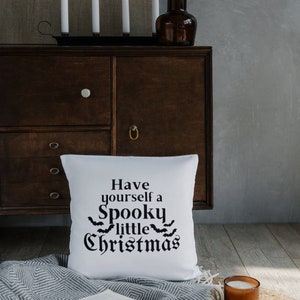 K&ouml;nnte beinhalten: Ein wei&szlig;es Kissen mit schwarzem Text, der "Have yourself a Spooky little Christmas" lautet, mit einer Reihe von Flederm&auml;usen &uuml;ber dem Wort "little". Das Kissen liegt auf einer grau-wei&szlig;en Decke mit einem Buch und einer Kerze.