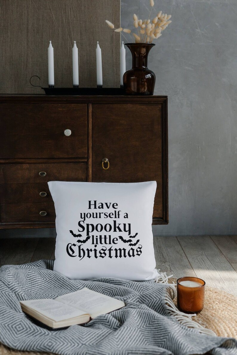 K&ouml;nnte beinhalten: Ein wei&szlig;es Kissen mit schwarzem Text, der "Have yourself a Spooky little Christmas" lautet, mit einer Reihe von Flederm&auml;usen &uuml;ber dem Wort "little". Das Kissen liegt auf einer grau-wei&szlig;en Decke mit einem Buch und einer Kerze.