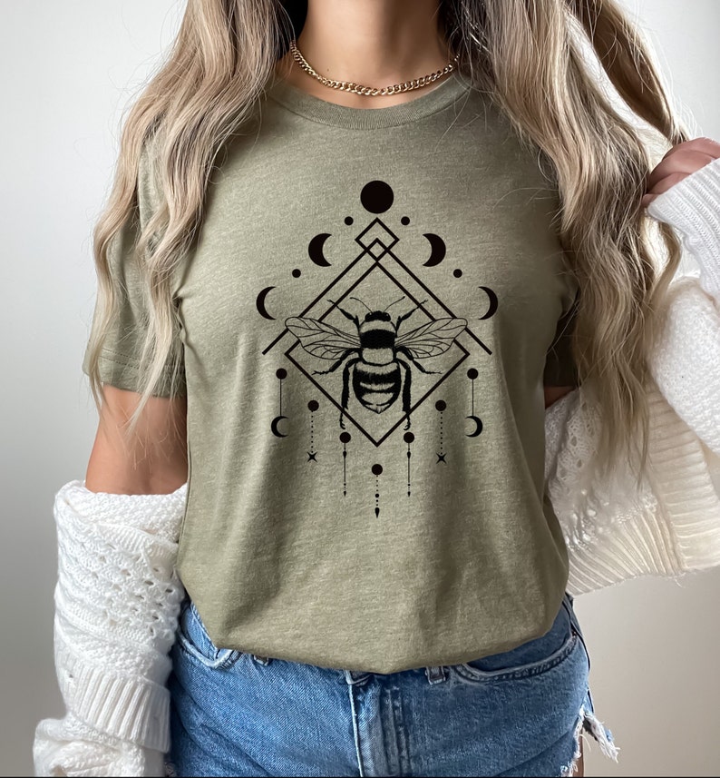 May include: A green t-shirt featuring a black graphic of a bee within a geometric design with moons and stars.