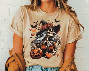 May include: A beige t-shirt featuring a graphic of a skeleton witch wearing a witch's hat adorned with flowers and holding a jack-o'-lantern. The witch is surrounded by bats and hearts.