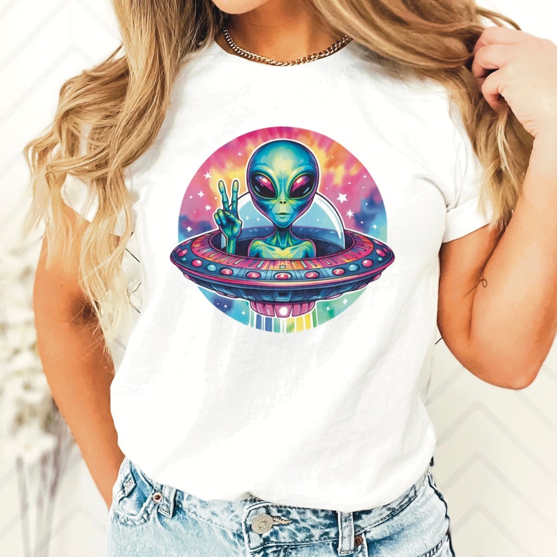 Puede incluir: Camiseta blanca con un dise&ntilde;o tie-dye que presenta un extraterrestre en un platillo volante haciendo un signo de paz.
