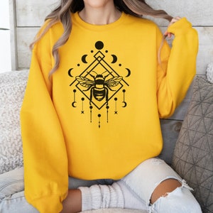 May include: A yellow sweatshirt featuring a black graphic design of a bee within a geometric shape, adorned with moons and stars.