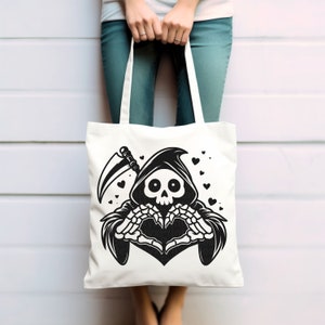 May include: A white tote bag with a black and white illustration of the Grim Reaper making a heart shape with its skeletal hands. The Reaper is wearing a hooded robe and holding a scythe. There are small black hearts scattered around the Reaper.