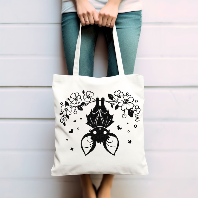 May include: A white tote bag with a black silhouette of a bat hanging from a branch with white flowers. The bat has a cute, cartoon-like appearance.
