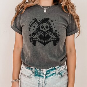 May include: A dark gray t-shirt with a black graphic of a skeleton wearing a hooded robe and making a heart shape with its hands. The skeleton is surrounded by small hearts.