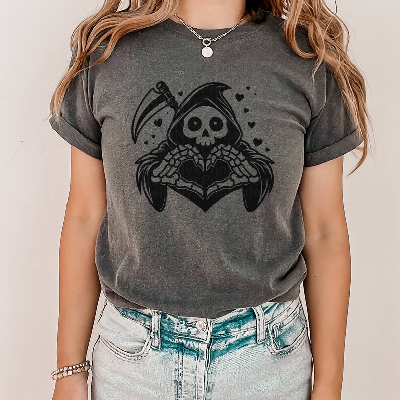 May include: A dark gray t-shirt with a black graphic of a skeleton wearing a hooded robe and making a heart shape with its hands. The skeleton is surrounded by small hearts.