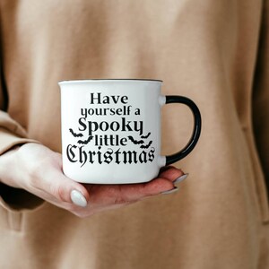 K&ouml;nnte beinhalten: Eine wei&szlig;e Keramiktasse mit schwarzem Henkel. Die Tasse hat einen schwarzen Text, der "Have yourself a Spooky little Christmas" lautet, mit einer Reihe von Flederm&auml;usen &uuml;ber dem Wort "little".