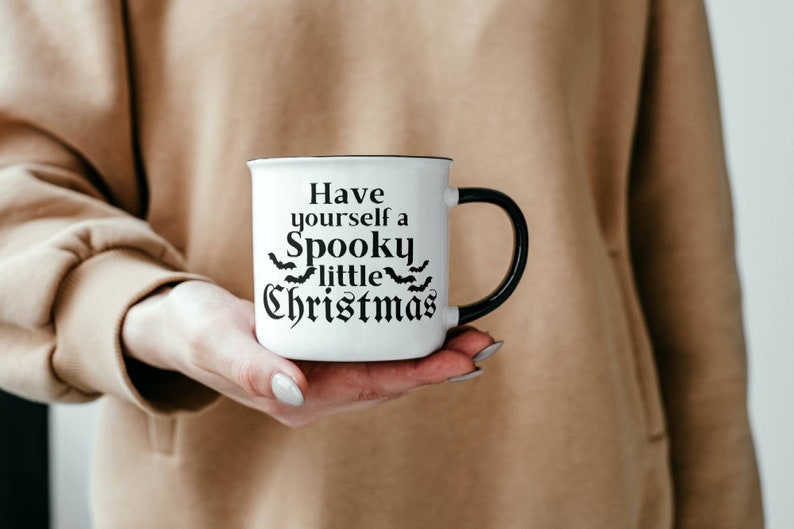 K&ouml;nnte beinhalten: Eine wei&szlig;e Keramiktasse mit schwarzem Henkel. Die Tasse hat einen schwarzen Text, der "Have yourself a Spooky little Christmas" lautet, mit einer Reihe von Flederm&auml;usen &uuml;ber dem Wort "little".