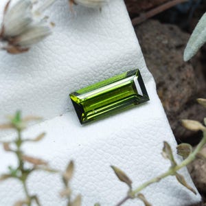 Può includere: Una pietra preziosa rettangolare con taglio baguette, caratterizzata da un colore verde oliva intenso e superfici sfaccettate che riflettono la luce. La pietra è posizionata su una superficie espositiva bianca e testurizzata, evidenziando la forma.