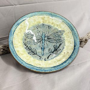 May include: A round ceramic plate featuring a central dragonfly illustration in dark blue ink. The plate has a cream-colored center and a textured blue rim. The design includes small leaf motifs flanking the dragonfly, with a glossy finish.
