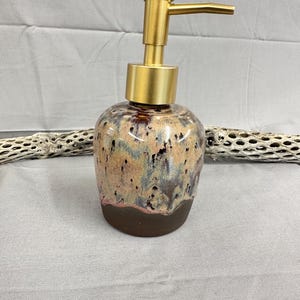 May include: A ceramic soap dispenser with a rounded body featuring a mottled glaze of tan, blue, and dark brown tones. The base is a solid dark brown, while the top is fitted with a brushed gold-toned metal pump mechanism for dispensing liquid.