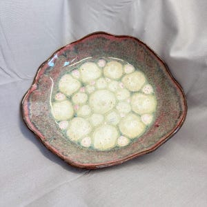 May include: A handmade ceramic bowl with a unique, organic shape. The bowl features a speckled, earthy color palette with a reddish-brown rim and a central design of cream-colored bubbles with pink accents. The bowl is approximately 8 inches in diameter.