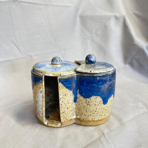 May include: A handmade ceramic condiment server with two compartments and matching lids. The stoneware piece features a speckled beige base with a vibrant blue glaze that drips down the sides. Each lid has a small, round knob.