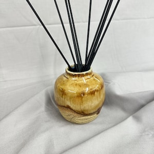 May include: A ceramic reed diffuser vessel with a glossy honey-brown glaze on the upper portion and a textured, speckled beige base. The round container holds a cluster of thin, straight black diffuser reeds extending outward from the opening.