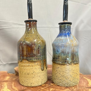 May include: Two ceramic oil bottles with black stoppers. One bottle has brown and green glaze, the other has blue and green glaze. Both bottles have a textured, tan base with a decorative pattern.
