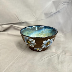 May include: A brown ceramic bowl with a blue and turquoise interior. The exterior features hand-painted white and light blue flowers with yellow centers. The bowl is a decorative piece for serving or display.