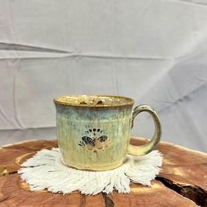 May include: A ceramic mug with a handle, featuring a butterfly and moon phase design. The mug has a speckled, glazed finish in shades of green and tan. It sits on a white macrame coaster, placed on a wooden surface.