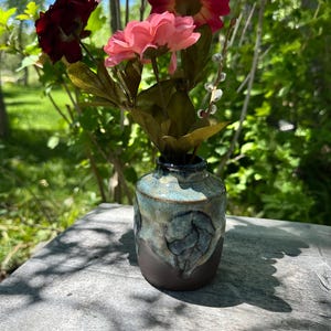 May include: A small ceramic vase with a dark brown base and a mottled blue and cream glazed top. The vase features a raised leaf-shaped relief on the front and contains a bouquet of artificial flowers in shades of pink and deep red.