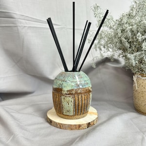 May include: A ceramic reed diffuser with a rounded shape, featuring a speckled light green glaze on the top half and a textured, brown vertical-striped pattern on the bottom half. Five thin black diffuser reeds are inserted into the opening.