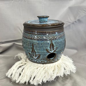 May include: A handmade ceramic garlic keeper with a lid. The jar is teal with brown accents and leaf-shaped cutouts. It sits on a white macrame coaster. The jar is approximately 6 inches tall and 5 inches wide.