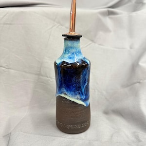 May include: A ceramic oil cruet with a cylindrical base in dark brown clay featuring a subtle embossed leaf pattern near the bottom. The upper portion is glazed in deep blue and white with a drip effect, topped with a metallic copper pour spout.