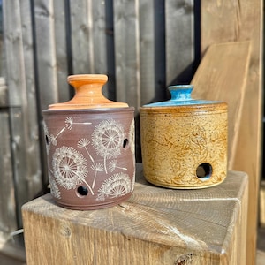 May include: Two cylindrical ceramic jars with lids. The left jar is dark brown with white dandelion seed head engravings and a light orange lid. The right jar is speckled tan with embossed leaf patterns and a bright blue lid.