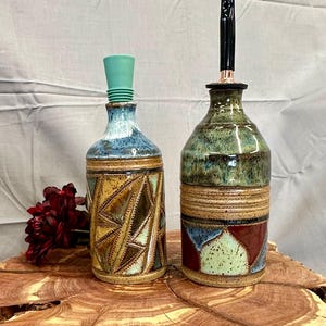 May include: Two decorative ceramic bottles with unique designs. One has a geometric pattern in earth tones and a teal stopper. The other has a green and brown glaze with a black and copper pour spout. Both are displayed on a wooden surface.