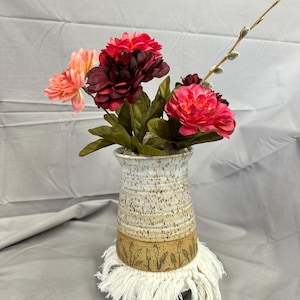 May include: A ceramic vase with a speckled white and tan finish, holding vibrant red, pink, and peach artificial flowers. The vase sits on a white macrame coaster with a wheat design.