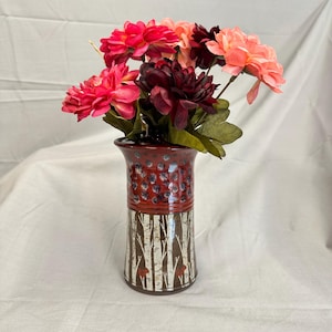 May include: A ceramic vase featuring a top section with a red glaze and dark blue spots, and a bottom section decorated with a white and brown birch tree pattern. The vase is filled with a bouquet of artificial flowers in shades of pink and dark red.