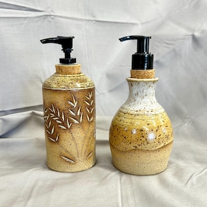 May include: Two ceramic soap dispensers with black pump tops. One is cylindrical with a wheat stalk design, the other is a rounded shape. Both are tan with speckled and textured finishes.