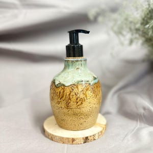 May include: A ceramic soap dispenser with a speckled tan base featuring an embossed leaf pattern and a smooth, light blue glazed neck. The dispenser is topped with a black plastic pump mechanism and rests on a circular, light-colored wooden coaster.