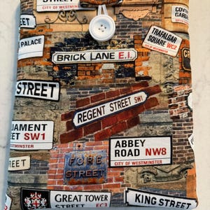 May include: A fabric book cover with a London street scene design. The cover displays street signs such as Oxford Street, Brick Lane, and Regent Street against a brick wall background. It has a button and loop closure.