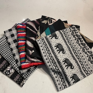 Puede incluir: Una colección de ocho bufandas de tela con diferentes patrones en negro, blanco, gris, rojo y verde. Las bufandas están dispuestas en forma de abanico sobre una superficie blanca.