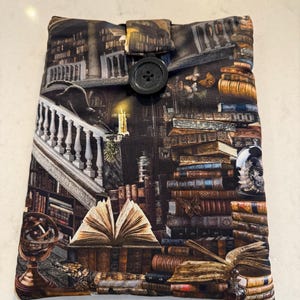 May include: A fabric book sleeve with a detailed print of a library interior, including stacks of books, a staircase, and a black cat. It has a button closure. The colour scheme features browns, tans, and blacks, offering protection for books.
