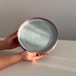 May include: A large, round, light blue ceramic serving bowl with a pink rim. The bowl is slightly textured and has a subtle, cloudy appearance.