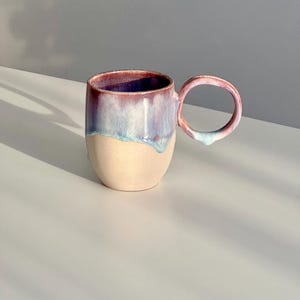 Peut inclure: Une tasse en céramique avec une poignée asymétrique unique. La tasse a une base beige clair avec un glaçage violet et bleu qui coule sur les côtés.